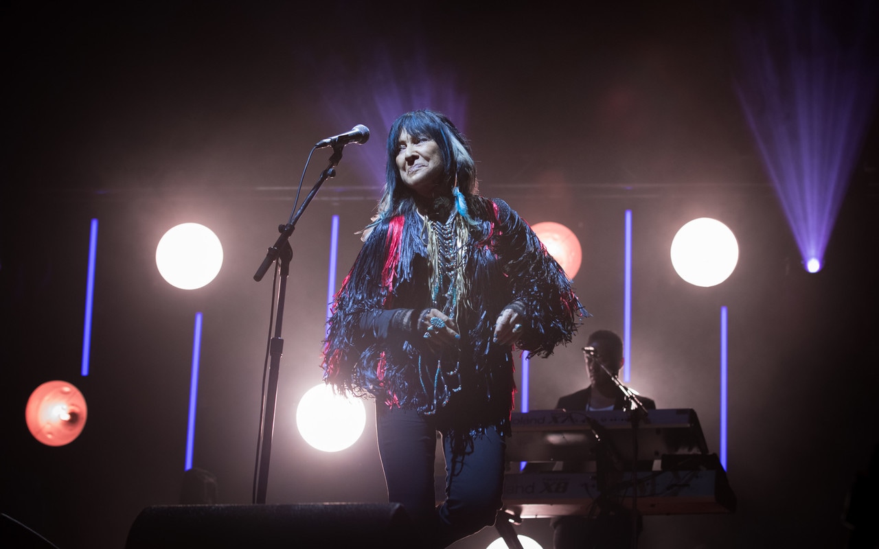 Woman wearing leather frayed tassel jacket, a beaded and boned necklace, and feathered earrings stands on a stage singing at a microphone. Five spotlight shine from behind her in the background.