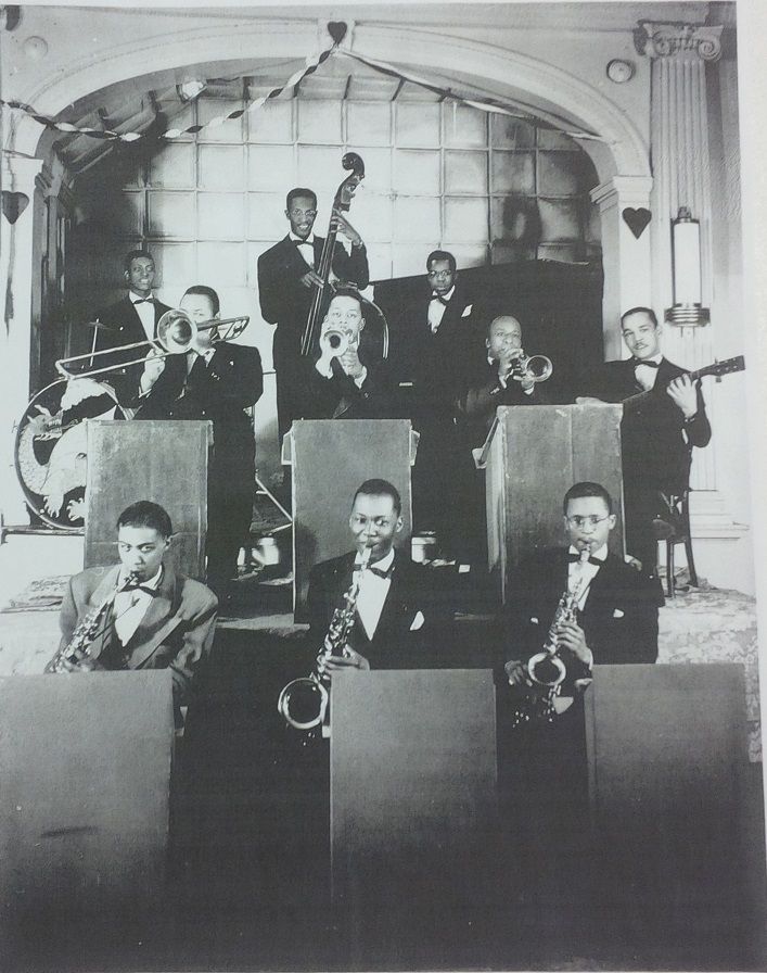  A band playing on stage. There are three brass instrument players in the front row. The second row has four musicians, three playing brass and one playing a string instrument. On stage is a pianist, bass player, and drummer.
