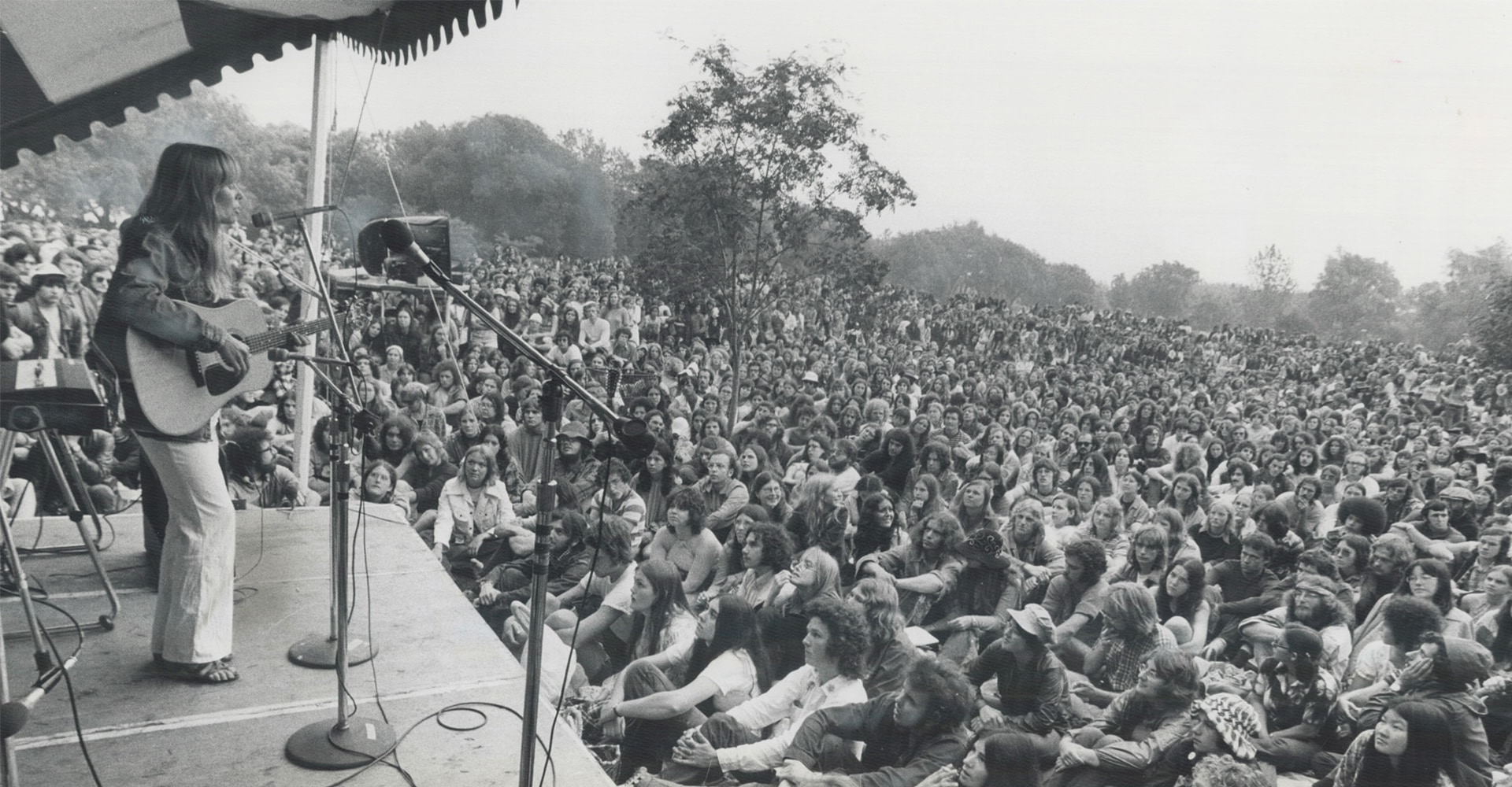 Une femme vêtue d’un chemisier et d’un pantalon tient une guitare debout sur une scène extérieure. Elle chante dans un microphone pendant que des centaines de personnes la regardent.