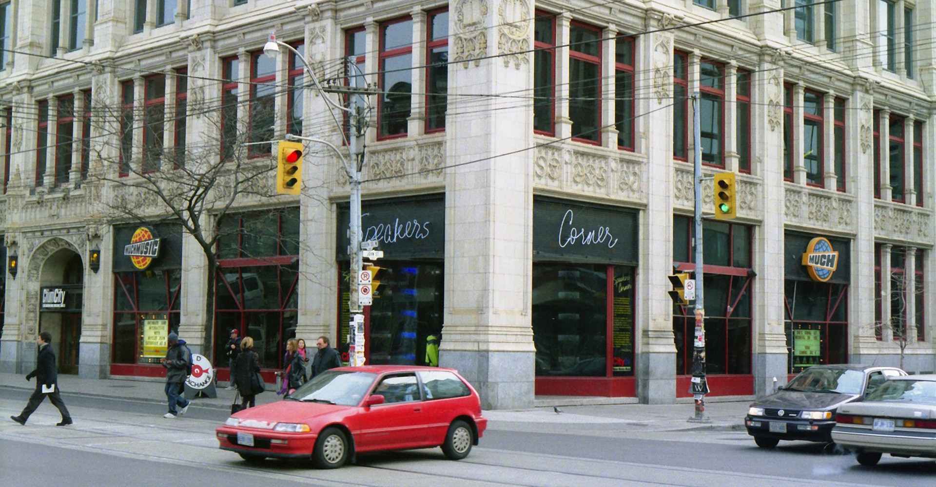 Exterior of a midrise building on the corner of an urban intersection. A car is driving through the intersection. Signs that read "Much" and "Speaker's Corner" are on the facade.