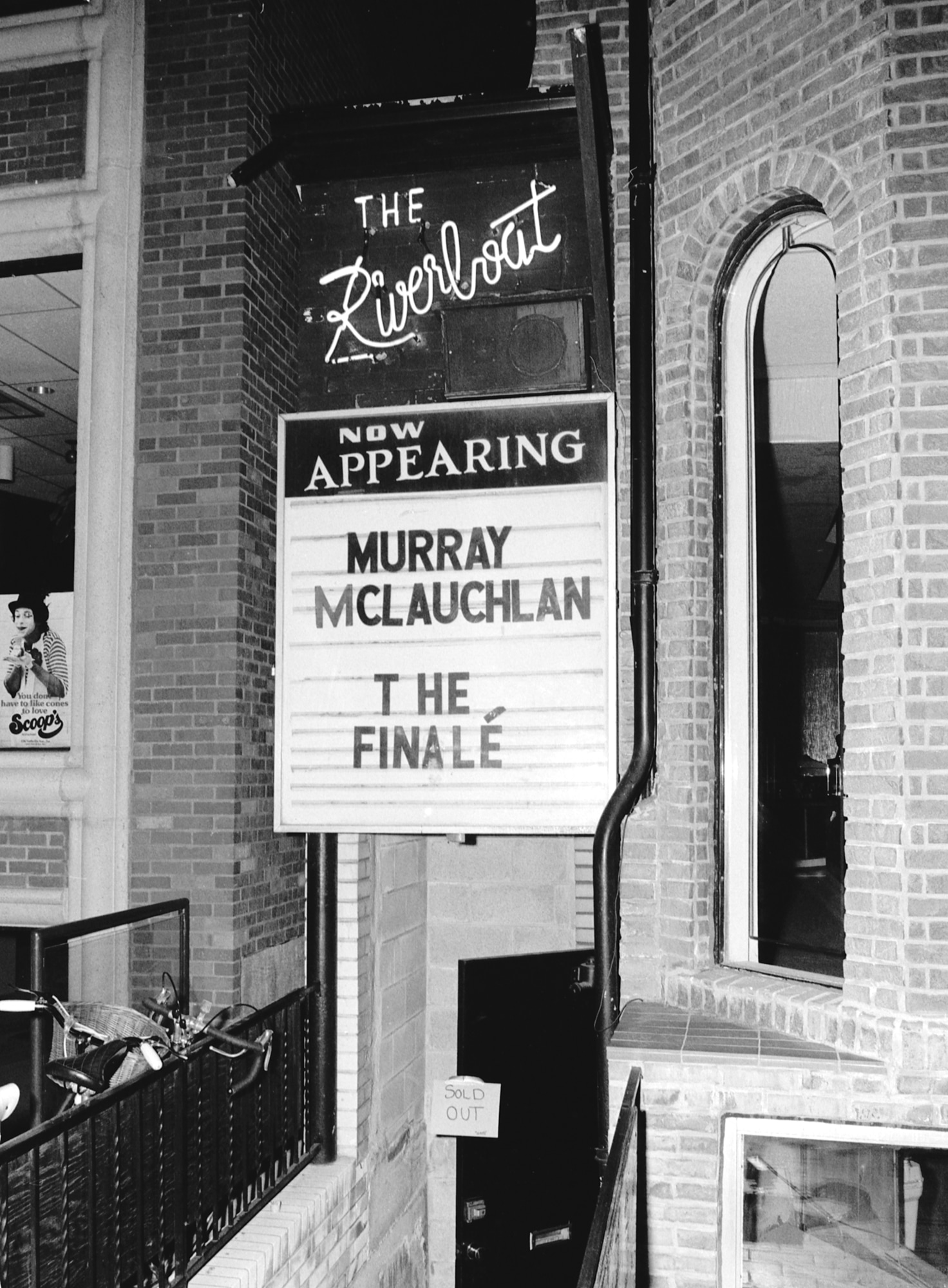  Outdoor brick building's entryway with stairs going down to the lower level. A sign above the door says " Riverboat. Now Appearing, Murray Maclaughlan, The Finale."