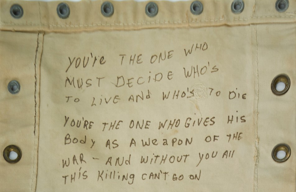 A canvas cot with words written on it in marker:  "You're the one who must decide who's to live and who's to die. You're the one who gives his body as a weapon of the war -- and without you all this killing can't go on."