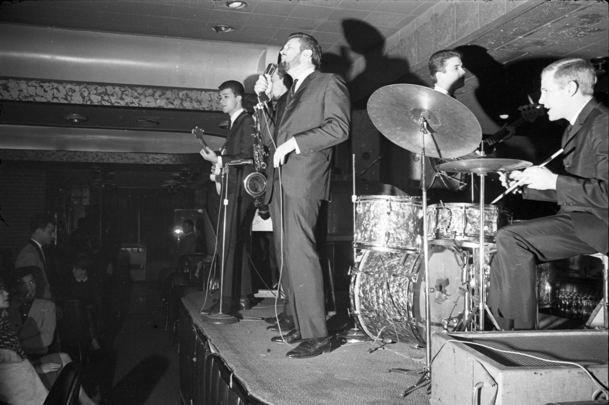 Cinq hommes en costume sur scène avec des instruments. On peut voir des spectateurs dans le coin inférieur gauche de l’image.