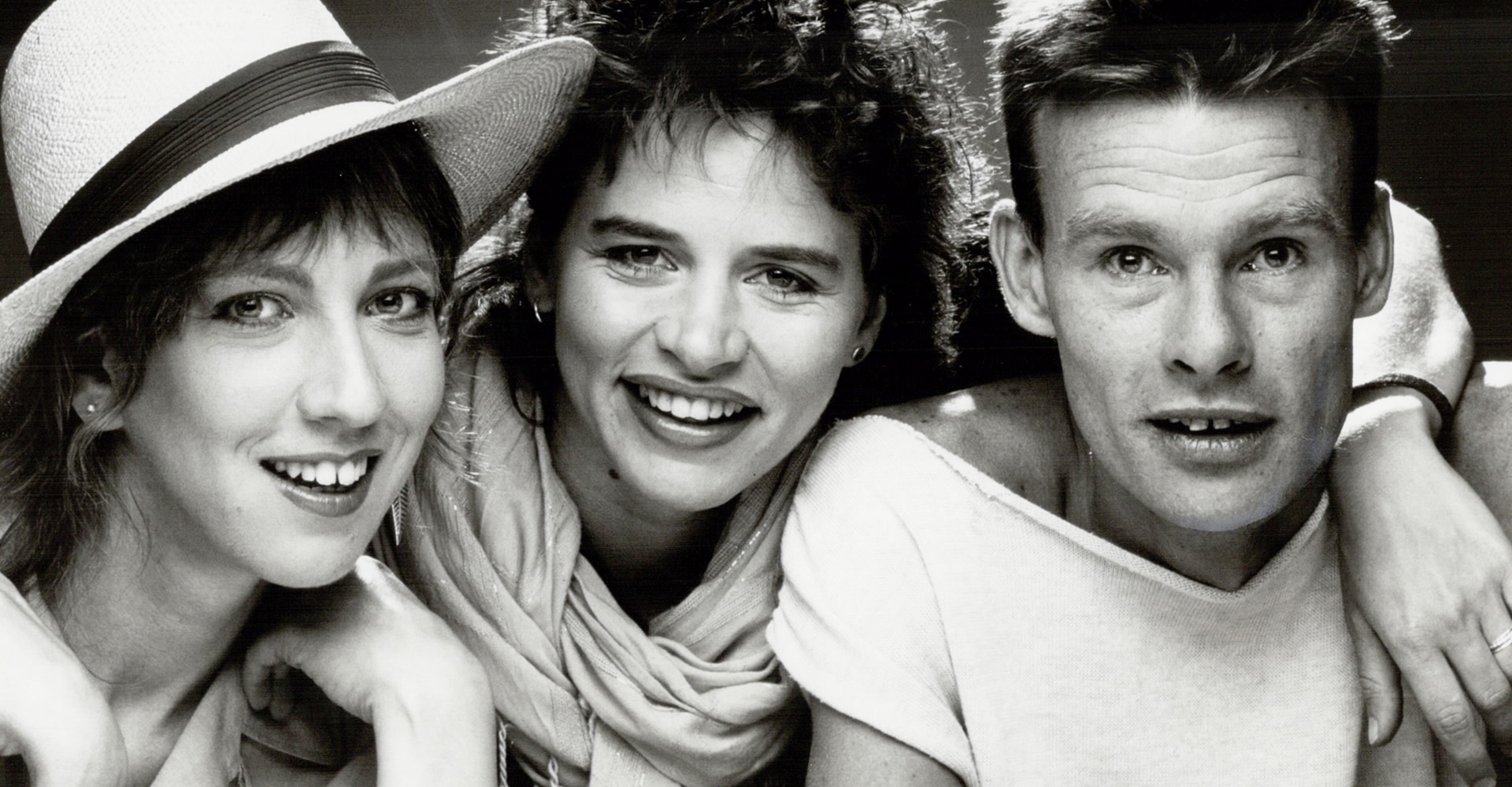 A group of three people, two women on the left, and a man at the right look into the camera. The woman on the left is wearing a hat. All have their mouths open in a slight smile.