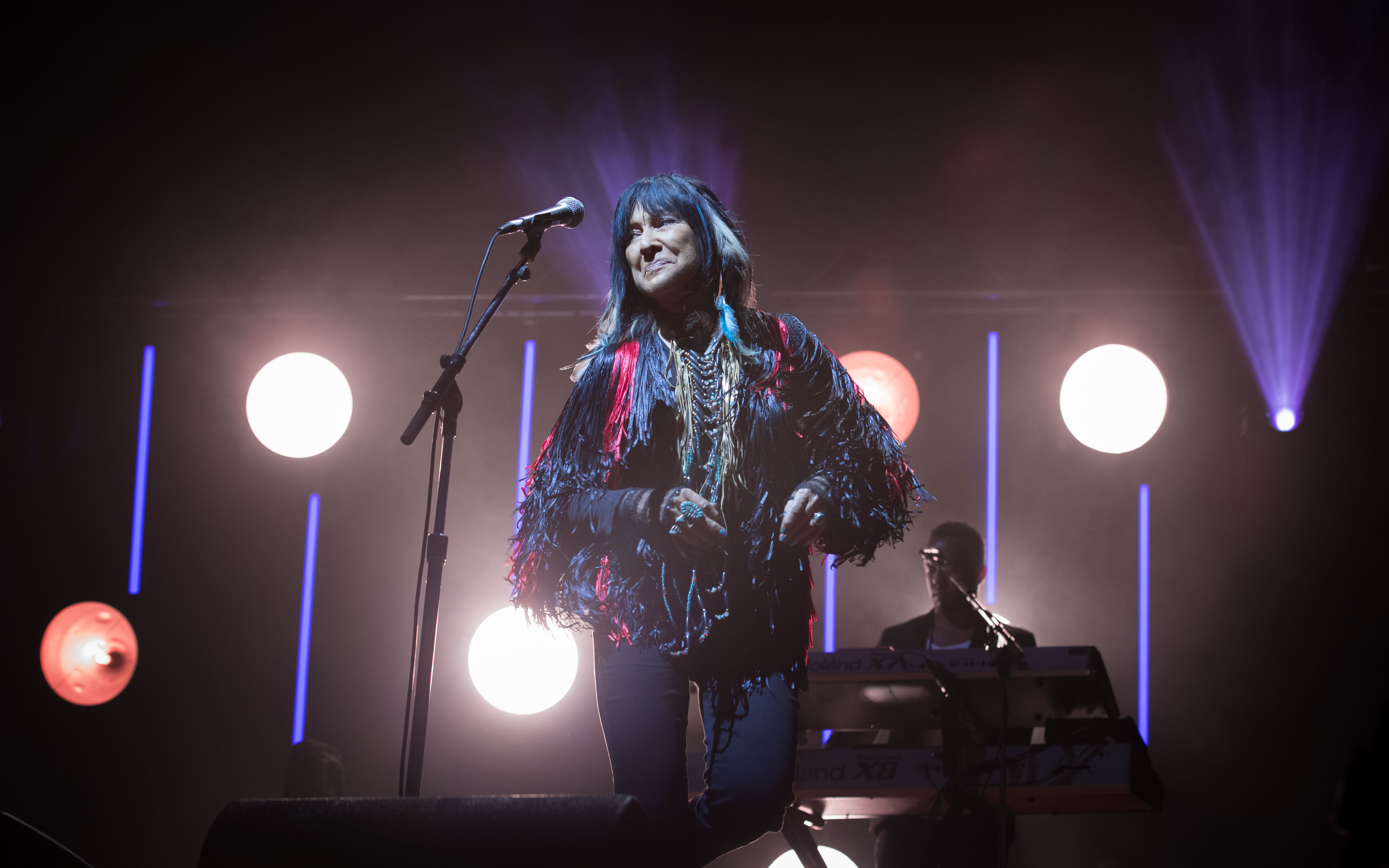 A woman with long hair, wearing a jacket with beads and fringe, stands on stage in front of a microphone