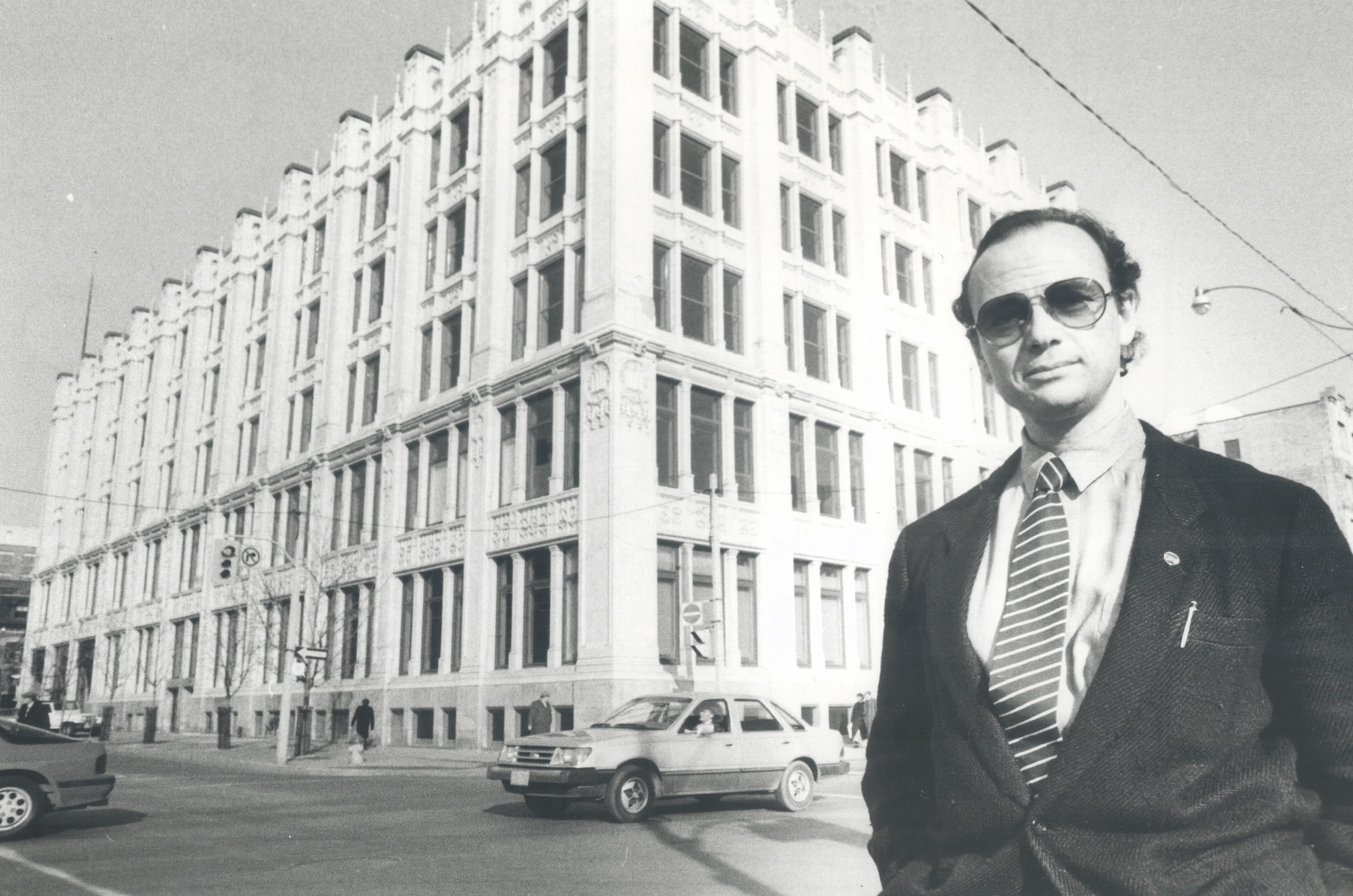 Un homme avec des lunettes de soleil et un complet se trouve sur un coin de rue. De l’autre côté de la rue se trouve un bâtiment orné de cinq étages.