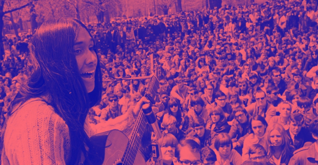 A woman with long hair playing acoustic guitar to a large outdoor crowd.