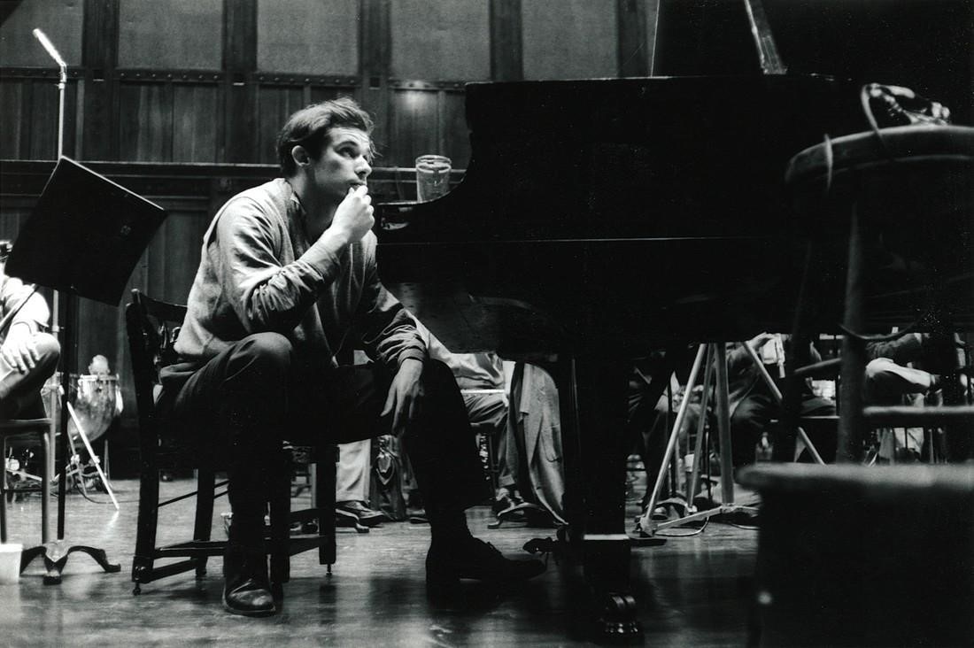 Un homme vêtu d’une chemise claire et d’un pantalon foncé est assis sur une petite chaise à côté d’un piano dans une salle de concert. Glenn Gould se touche le menton et se trouve dans un état contemplatif. Les membres d’un orchestre sont visibles à l’arrière-plan.