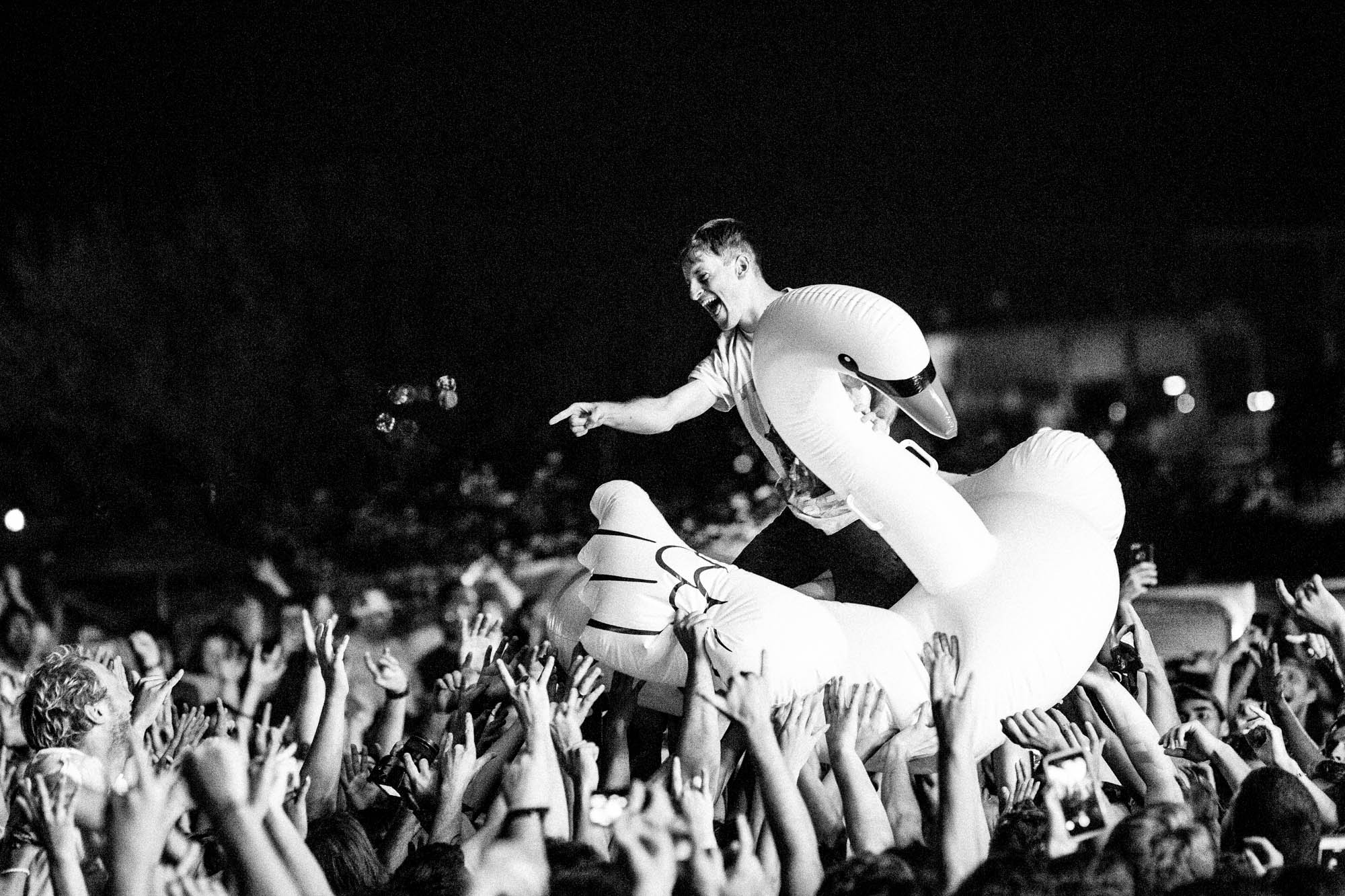 Stefan Babcock rides a large inflatable white duck through a concert crowd on Echo Beach in Toronto, held aloft by hundreds of hands. He is dressed casually in a white graphic t-shirt. He points outward to the audience while smiling.