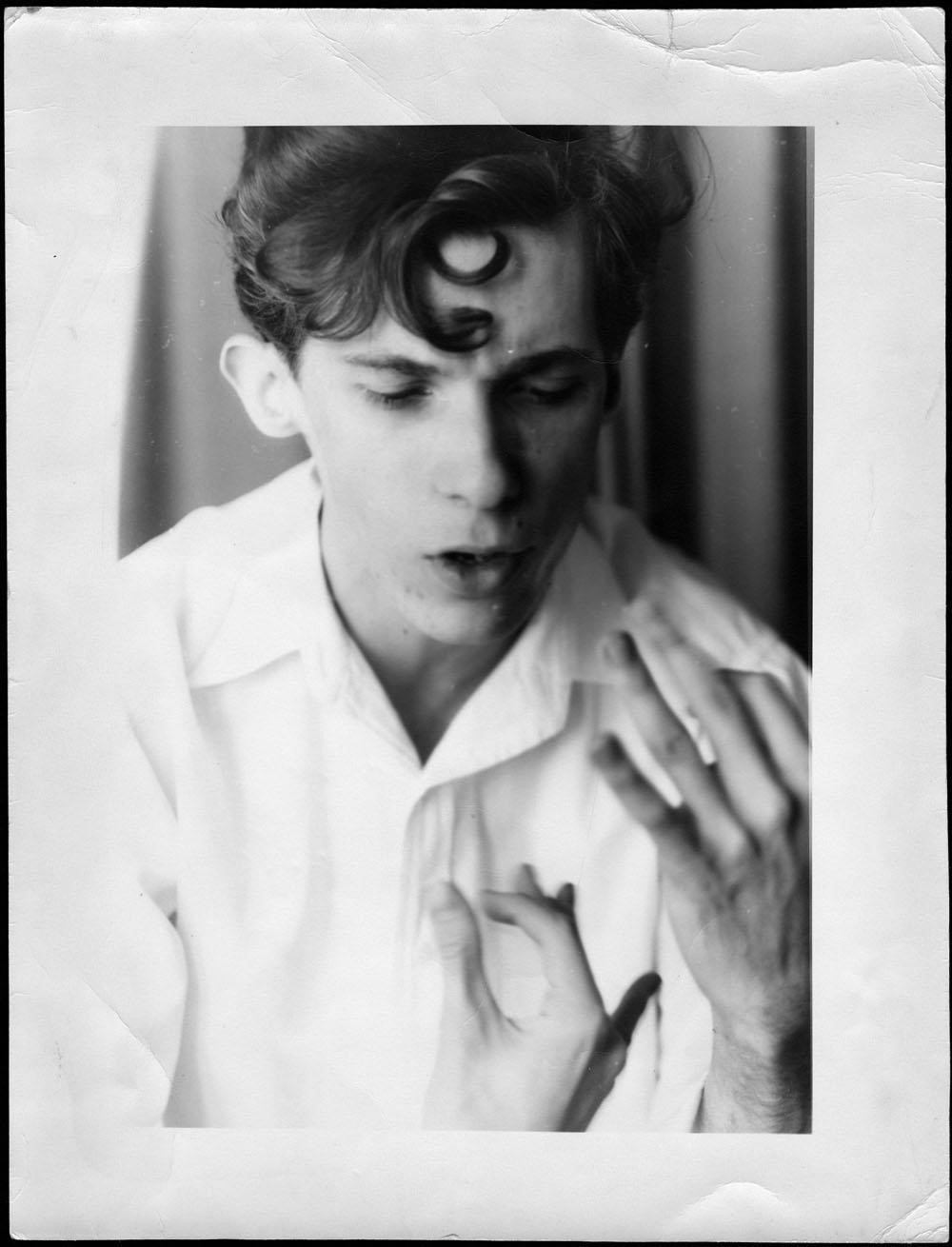 Portrait of a young Glenn Gould dressed in a white shirt. Gould is looking slightly down while speaking. He holds his hands in a gesture in front of his body.