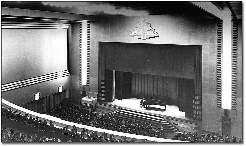 A black and white image looking down to a stage from a balcony. There are rows of seats leading up to the stage. A grand piano sits on the stage. Stairs lead up to the stage.