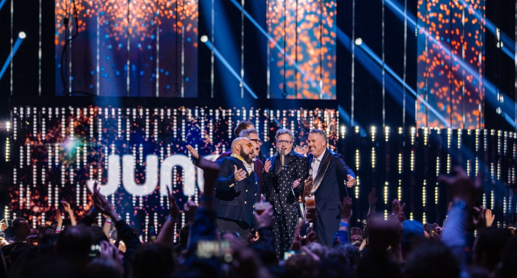: A group of men in variously coloured blue suits smile and sing into a microphone on stage. The man on the right has a guitar on a strap. Behind them is a lit sign that reads “JUNOS”. In front of them, a large audience waves their hands and their smartphones.Photo by Ryan Bolton.