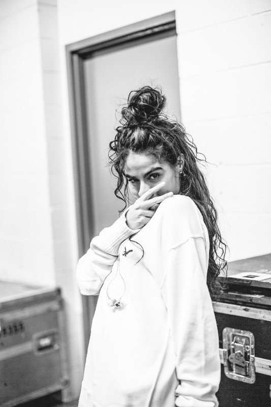 In a black and white photo, Jessie Reyez shows the peace sign with her right hand, which is held in front of her face, while backstage at the JUNO Awards. Her hair is up in a messy bun and she is wearing a baggy white sweatshirt. 