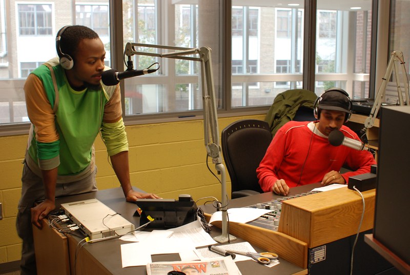 A colour photograph featuring two men in a radio room. Both are in front of microphones and are wearing headphones. In front of them a radio mixing console sits on a desk along with papers. 