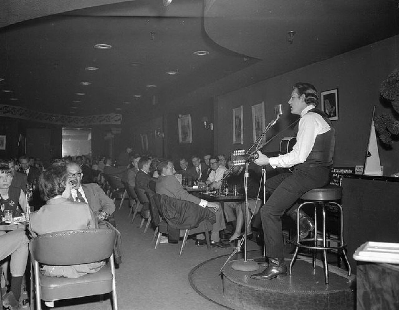 A black and white image of a man sitting on a stool on a small stage in the right hand corner, playing an acoustic guitar and singing into a microphone. He is wearing a vest, dress pants, and dress shoes. The audience of men and women in various dress clothes are to the left of the singer. The ceiling is low and dark and the room is smoky.
