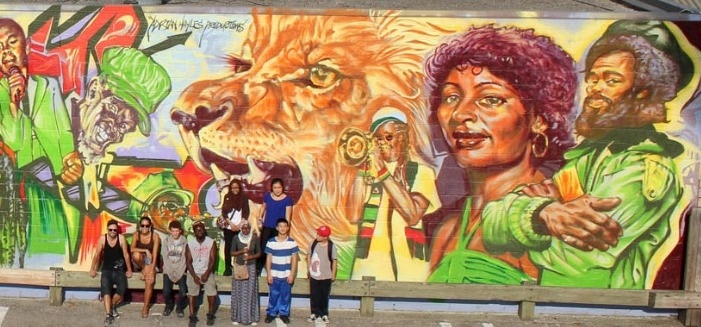 A group of nine artists stand in front of a large-scale mural painted on the back of commercial buildings. The mural backs onto a parking lot and depicts over a dozen different musicians, as well as a lion. It is painted in vibrant colours, mostly red, yellow and green.