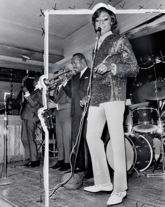A singer in white pants and sequinned jacket holds a microphone on stage. A trumpet and saxophone player stand next to them. Behind them is a drum kit.