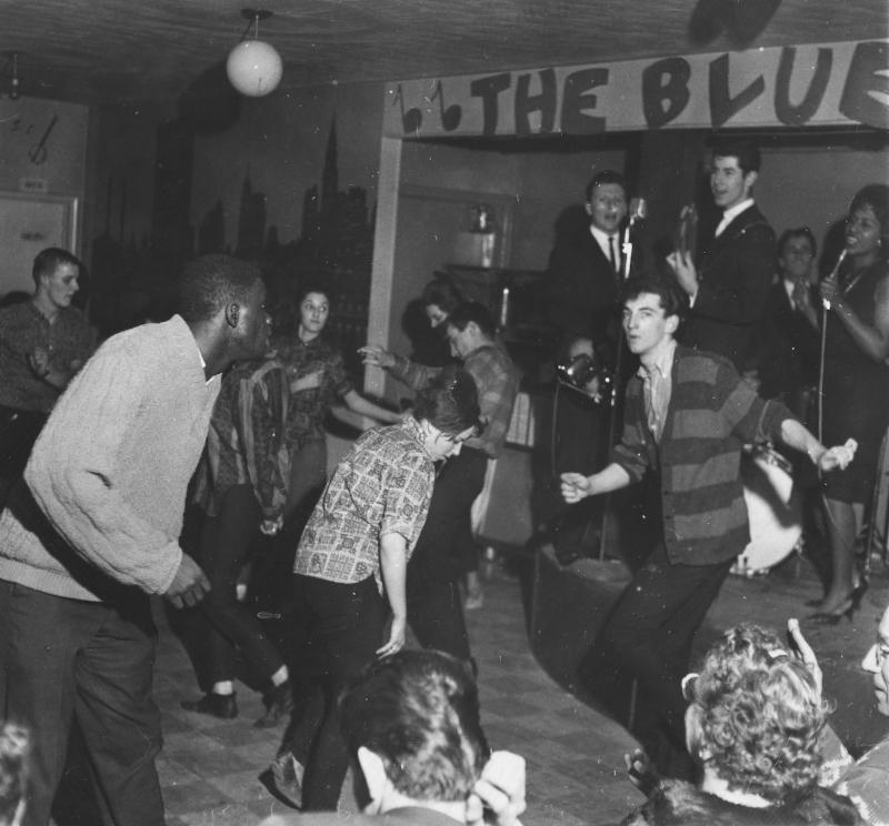 Une photo en noir et blanc d’un groupe jouant sur une scène surélevée. En dessous de la scène et devant celle-ci, plusieurs personnes habillées de façon informelle dansent et regardent les artistes.