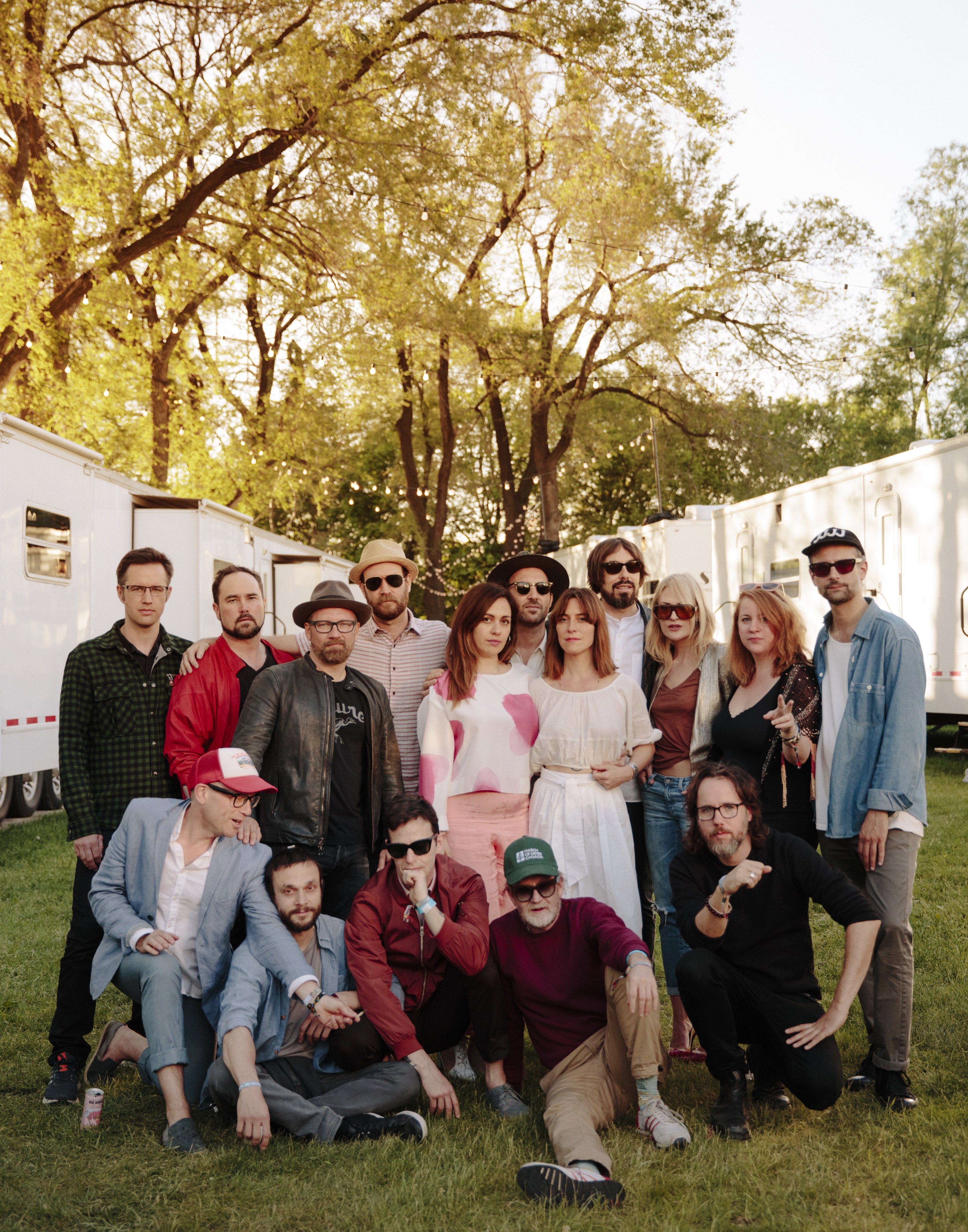 16 people stand on grass on a summery day; five sit or kneel, while 11 stand behind in a variety of poses, all looking at the camera. A mix of men and women, they are informally dressed. The group stands between two long rows of white trailers. 