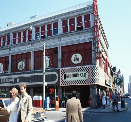  A colour photograph of the exterior of a three-storey building which is located at the corner of two streets. On the ground floor of the building signs advertise the “Hard Rock Cafe”. Above, on the second and third floors is a stone facade, outside of which hangs a sign advertising “Nickelodeon.”