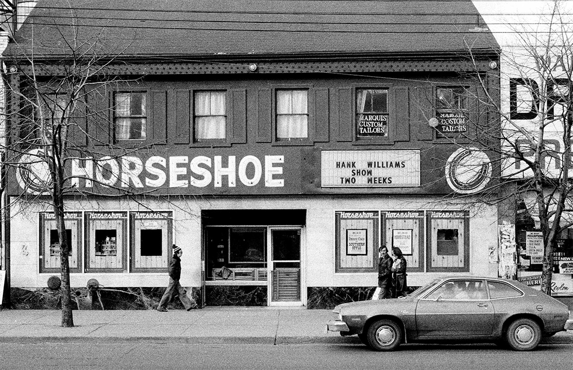 An exterior black and white photo of a two-story building. A large sign affixed to the left-hand side of the building reads: Horseshoe. On the right, a marquee announces upcoming acts of Hank Williams Show Two Weeks. A 1970s-era car is parked out front. Photo by Patrick Cummins.