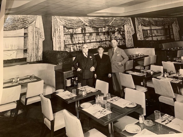 A black and white photograph of the interior of a restaurant. Numerous tables set up for restaurant service can be seen, with wide booth seats towards the back. In the middle of the frame stand three men, all wearing suits. 