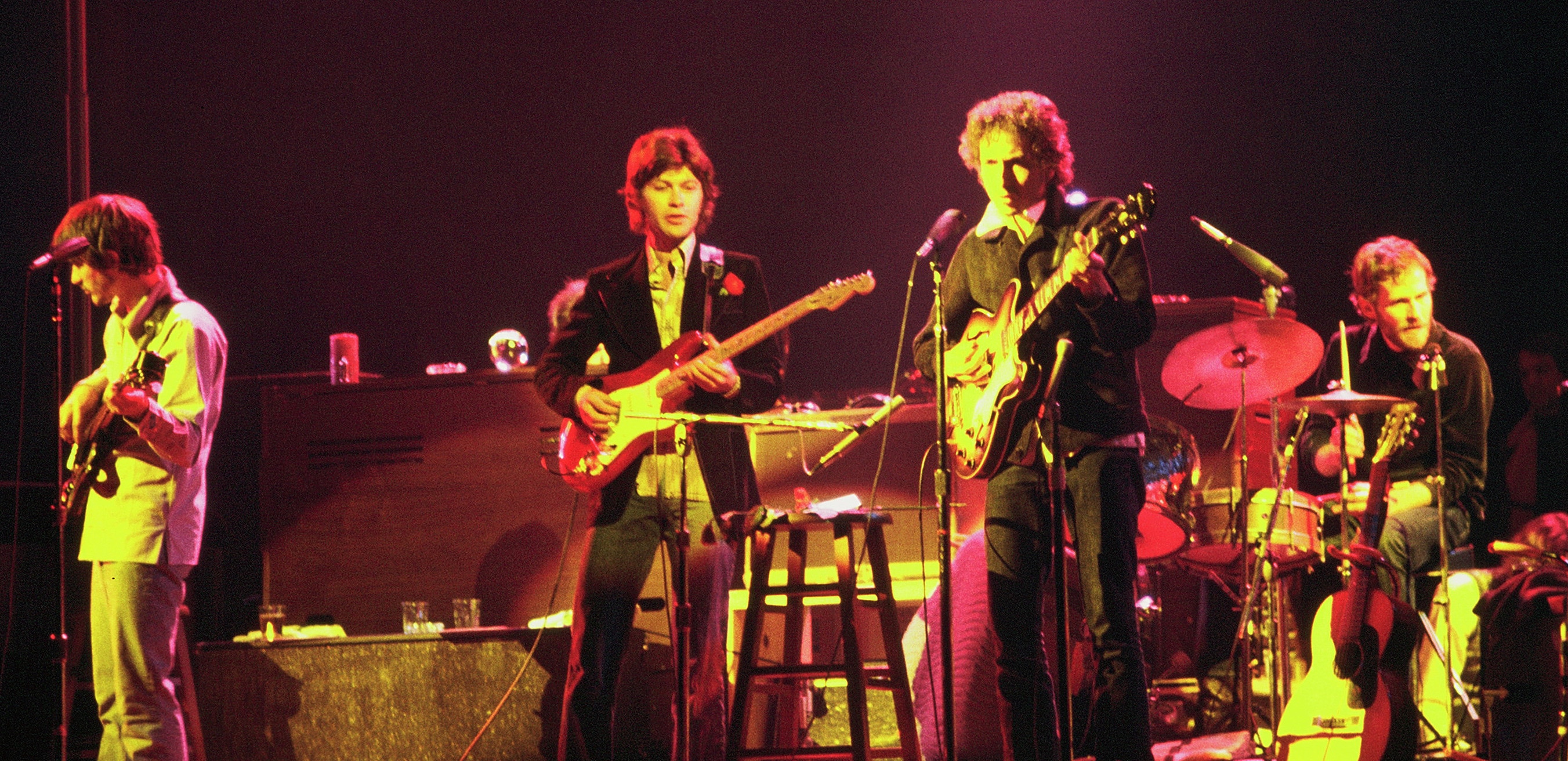 Four men stand on stage dressed in 1970s clothes. One holds a guitar and stands in front of a microphone. To his left two men hold and play guitars. On the far right, a man sits at a drum kit playing.