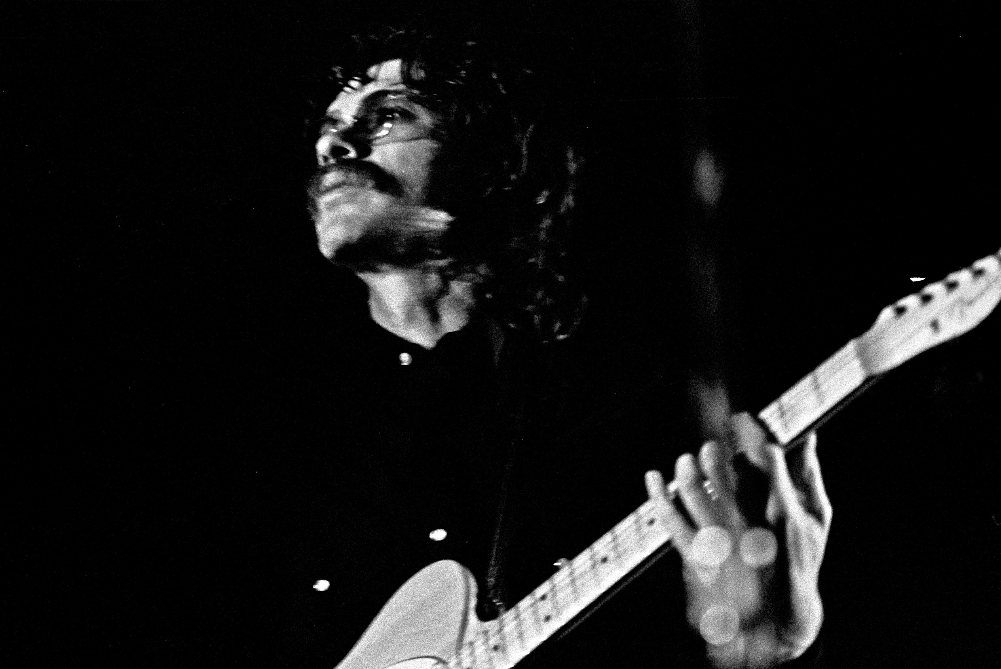 Une photo en noir et blanc d’un homme jouant de la guitare. Il porte des lunettes et a une moustache. Il détourne le regard de l’objectif et regarde droit devant lui. 