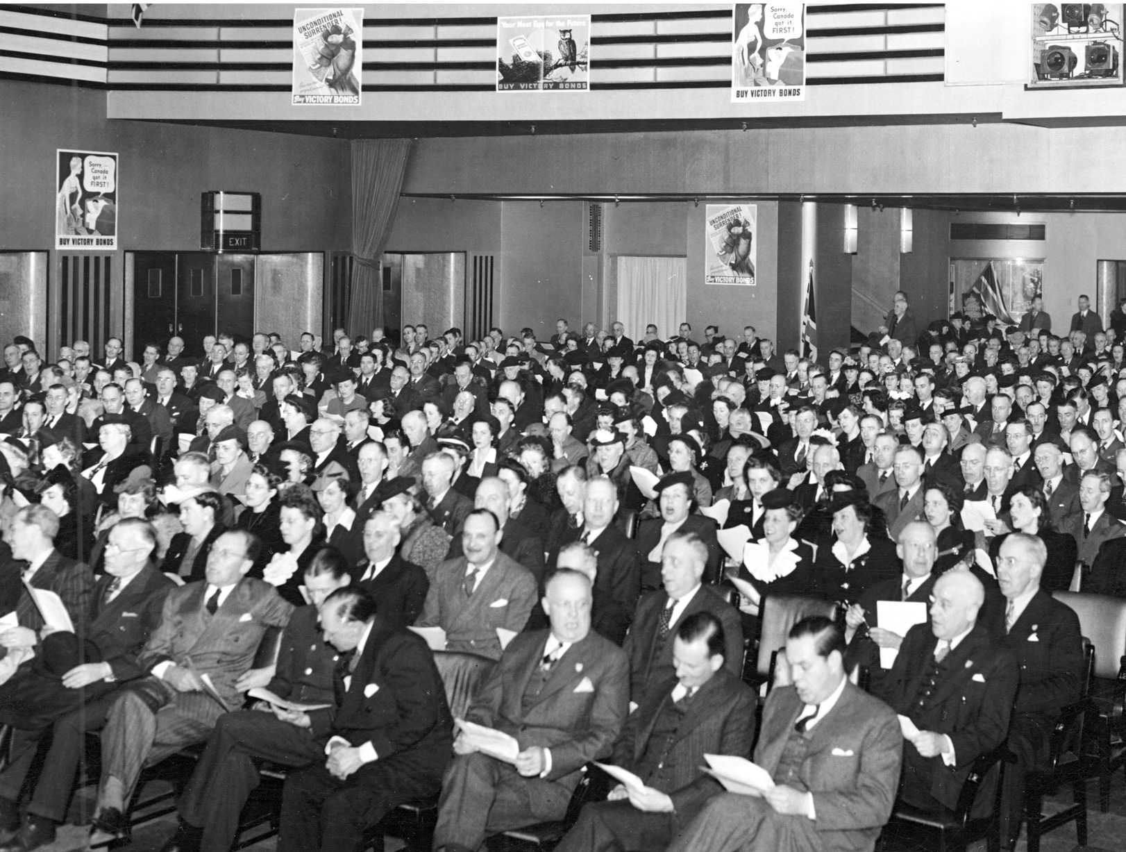 Black and white photo of an audience sitting in a theatre featuring Art Deco decoration. Every seat is full. Photo dates to 1943.
